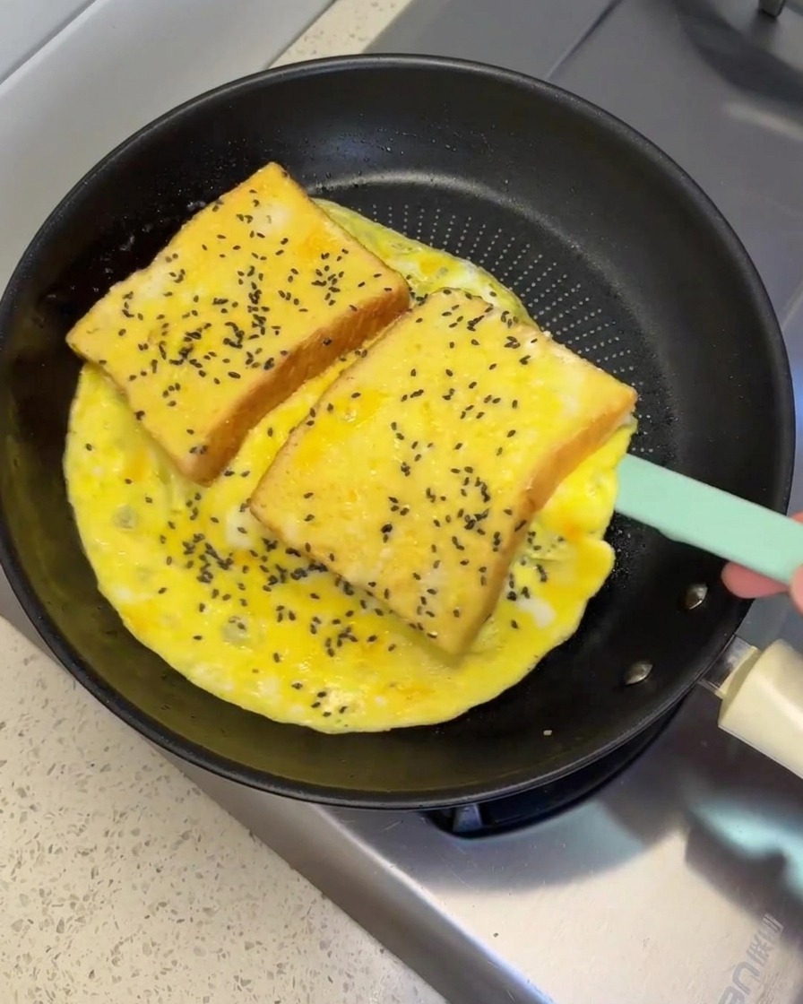Une spatule en silicone soulevant et retournant une grande omelette aux œufs contenant deux tranches de pain dans une poêle.