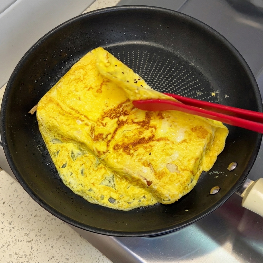 Des pinces en silicone rouge repliant les bords qui dépassent dune omelette aux œufs cuite sur deux tranches de pain dans une poêle.