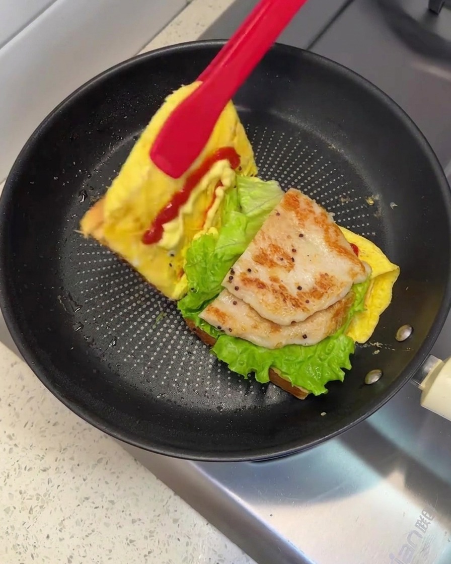 Une paire de pinces rouges soulevant et repliant un côté du pain pour fermer le sandwich dans la poêle.