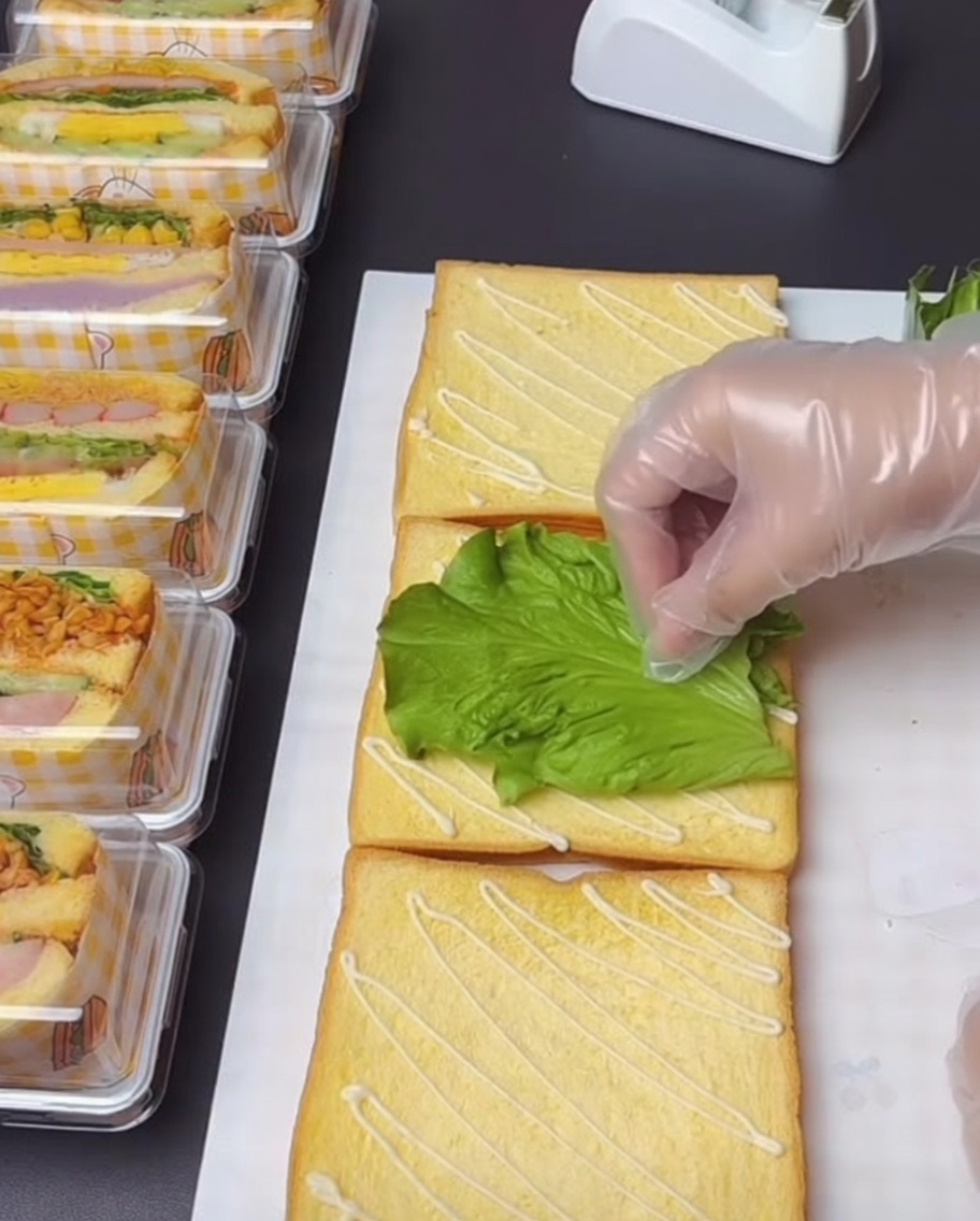 Une main gantée déposant délicatement une feuille de laitue verte et fraîche sur une tranche de pain nappée de sauce.