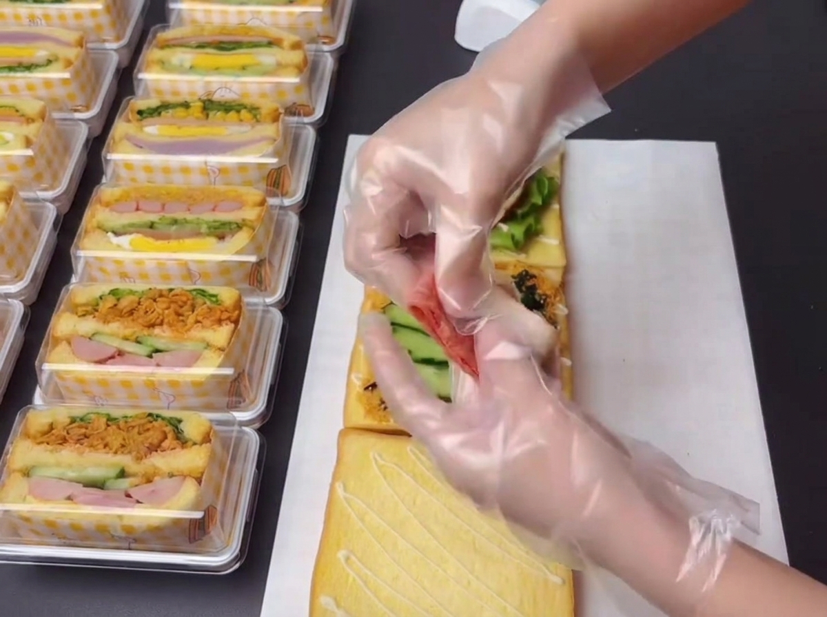 Des mains gantées tenant et disposant de petits morceaux de bacon cuit sur la structure du sandwich.