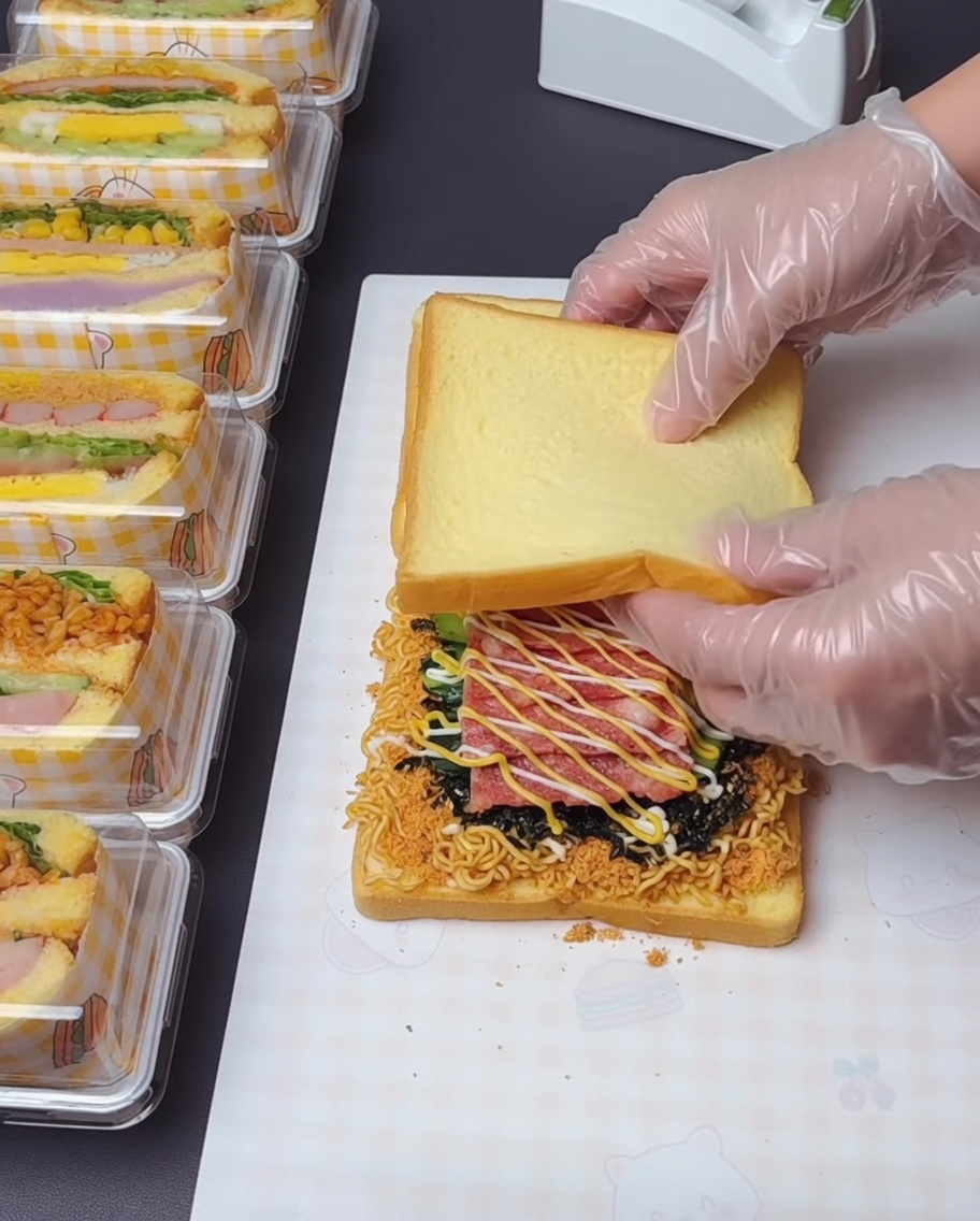 Une paire de mains gantées soulevant et superposant la tranche supérieure de pain sur les couches garnies en dessous.