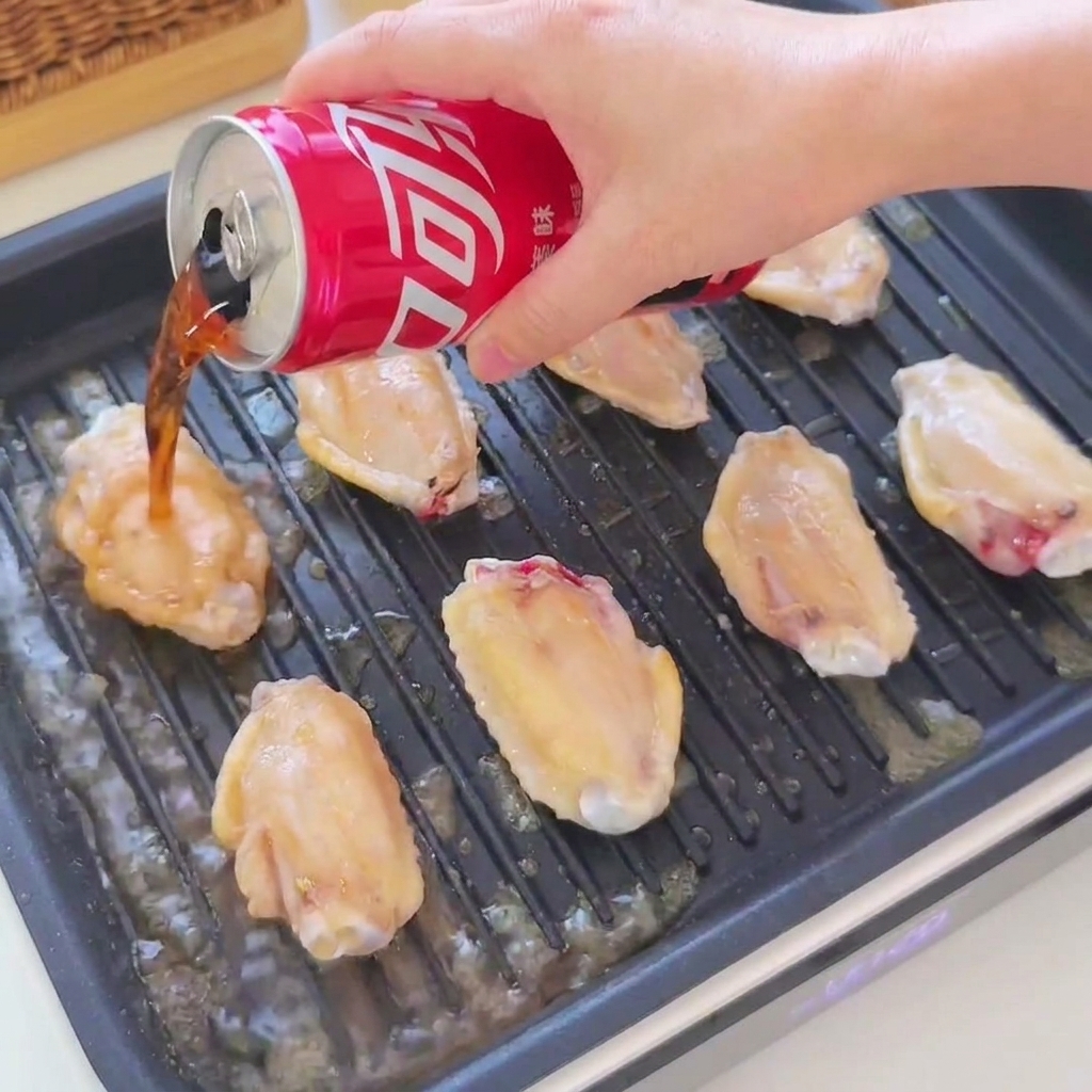 Cola et sauce soja versés dune canette rouge sur des ailes de poulet dorées grésillant dans un gril noir.