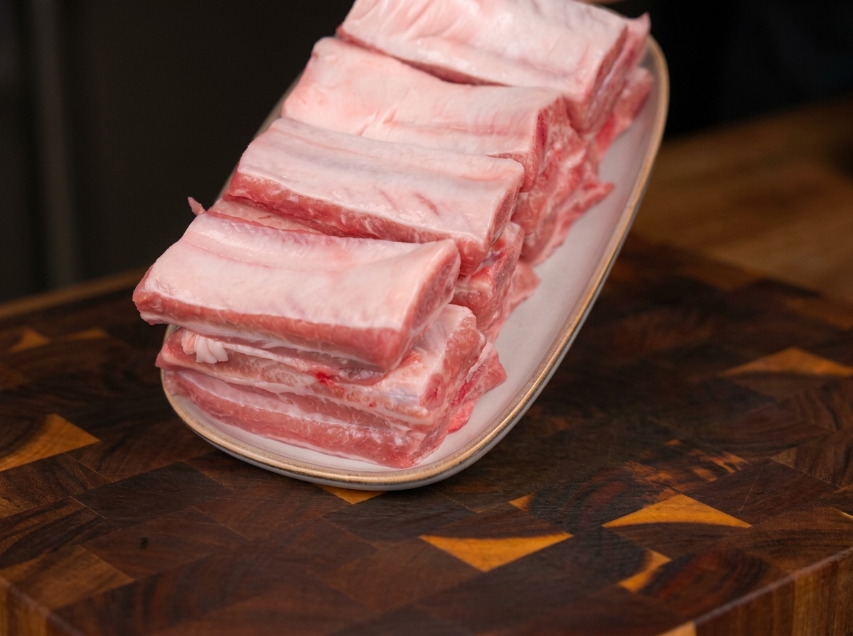 Travers de porc crus et frais, soigneusement empilés sur une assiette blanche ovale posée sur une planche à découper en bois.