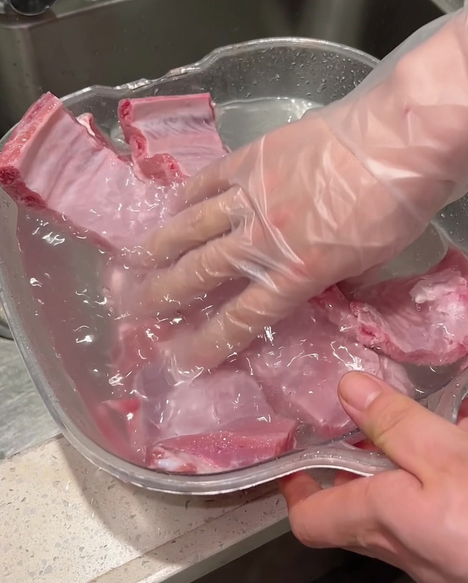 Des mains gantées frottant et lavant des travers de porc crus dans un récipient rempli deau.
