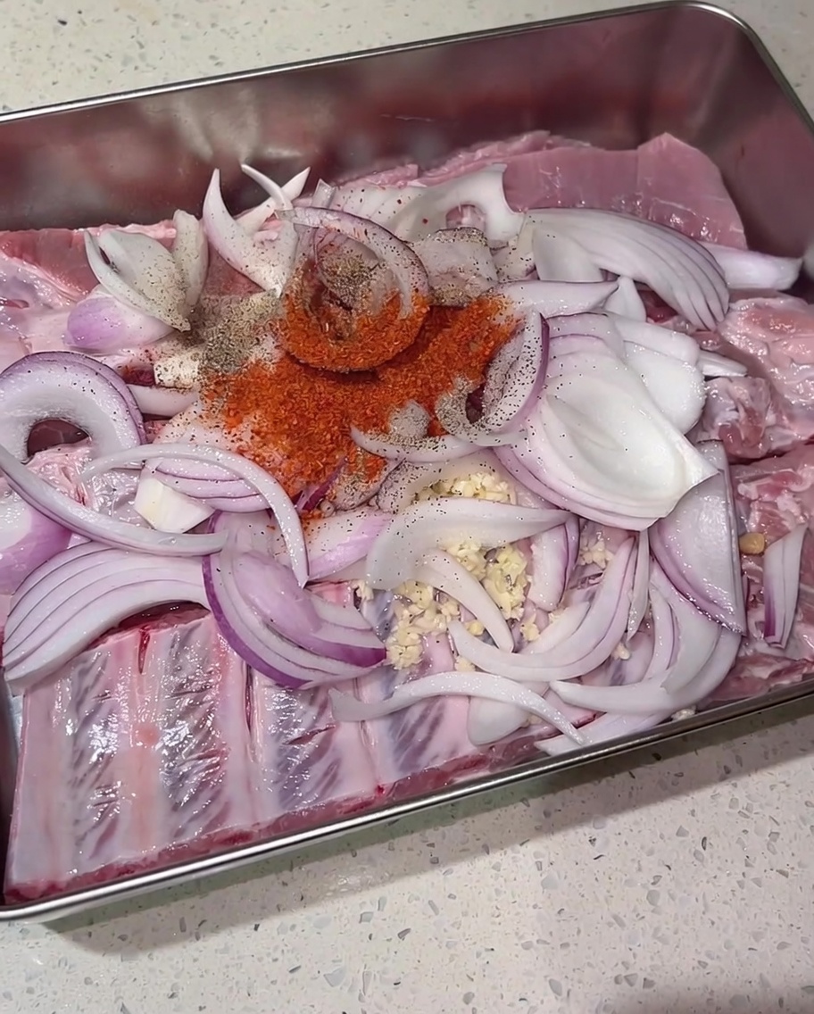 Un tas de piment rouge en poudre, de poivre noir et de sucre roux reposant sur des travers de porc crus et des oignons rouges tranchés dans un plateau en métal.