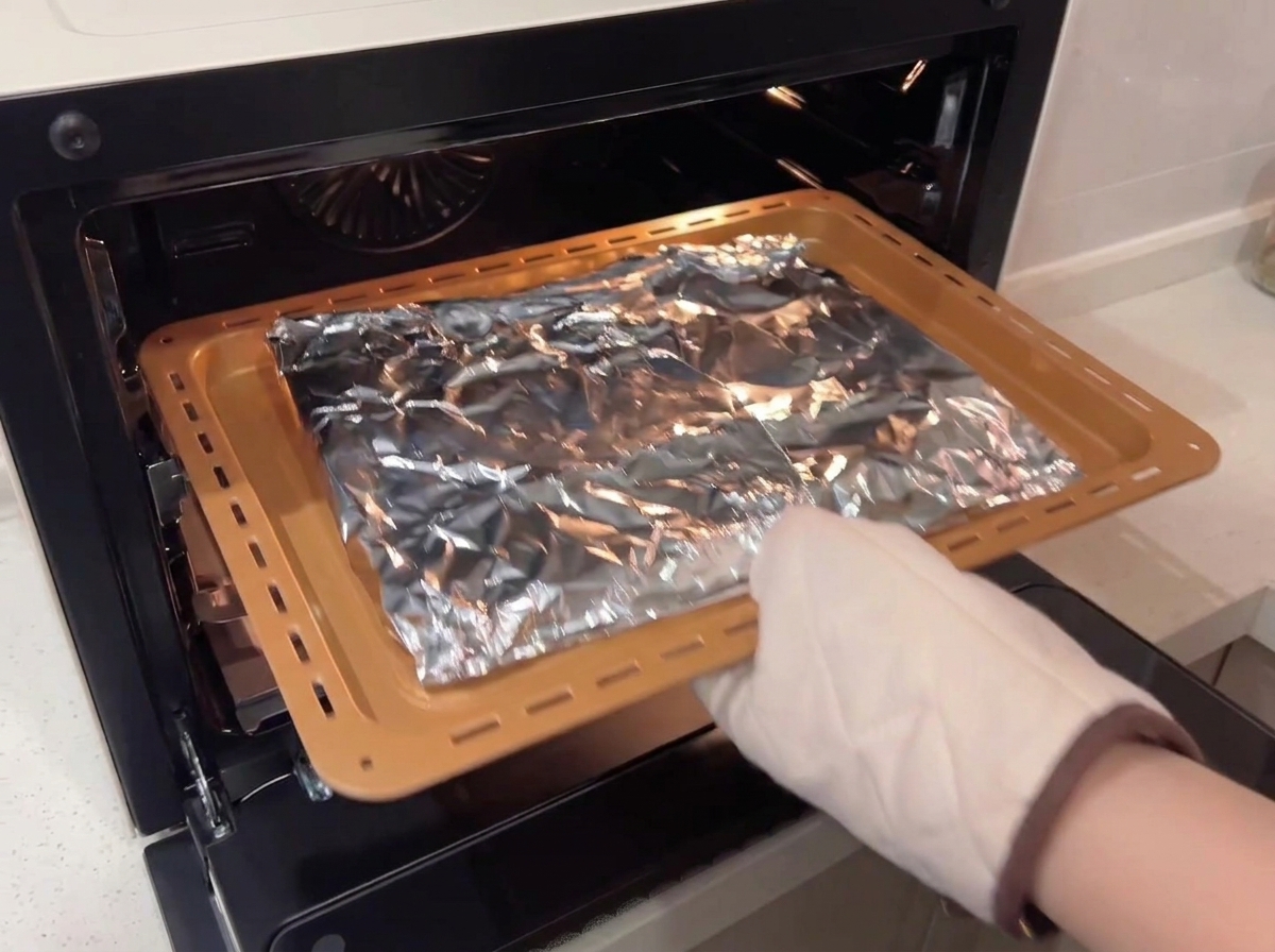 Une personne portant un gant de cuisine de protection plaçant une plaque de cuisson avec des côtes enveloppées de papier aluminium dans un four à convection.