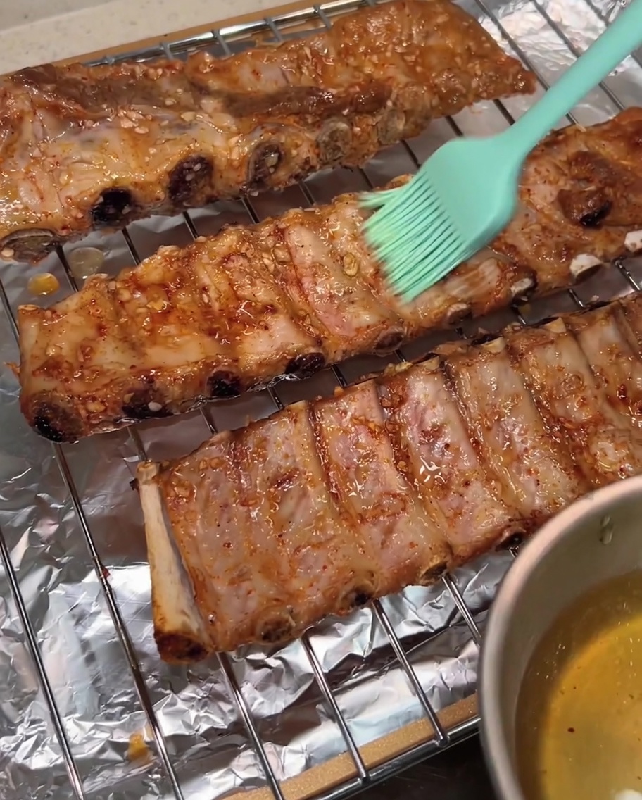 Un pinceau en silicone vert appliquant un revêtement brillant deau de miel à la surface des travers de porc rôtis sur une grille.
