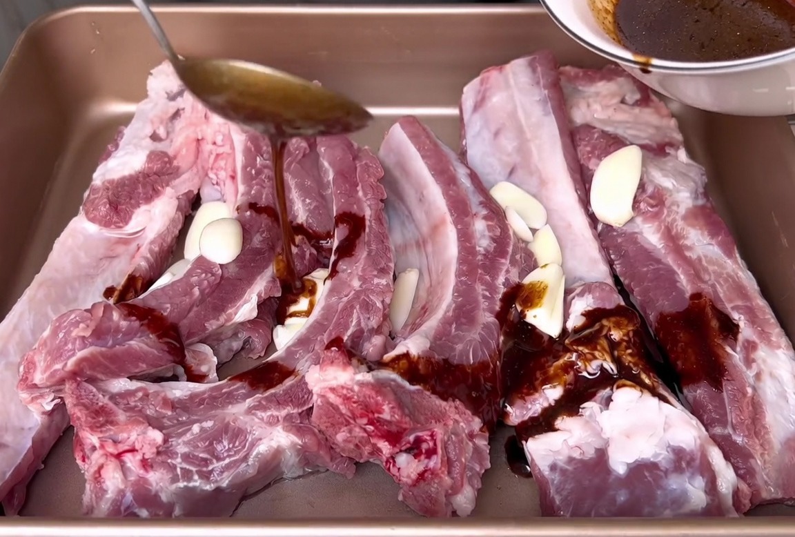 Versement dune marinade à base de sauce soja depuis un bol blanc sur des côtes de porc crues et des tranches dail dans un plat de cuisson.