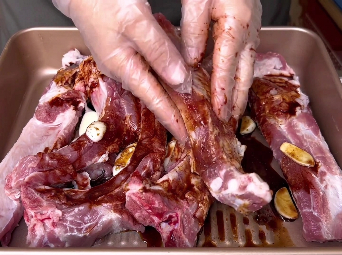 Des mains gantées frottant vigoureusement une marinade liquide sombre dans des côtes de porc crues à lintérieur dun plateau doré.