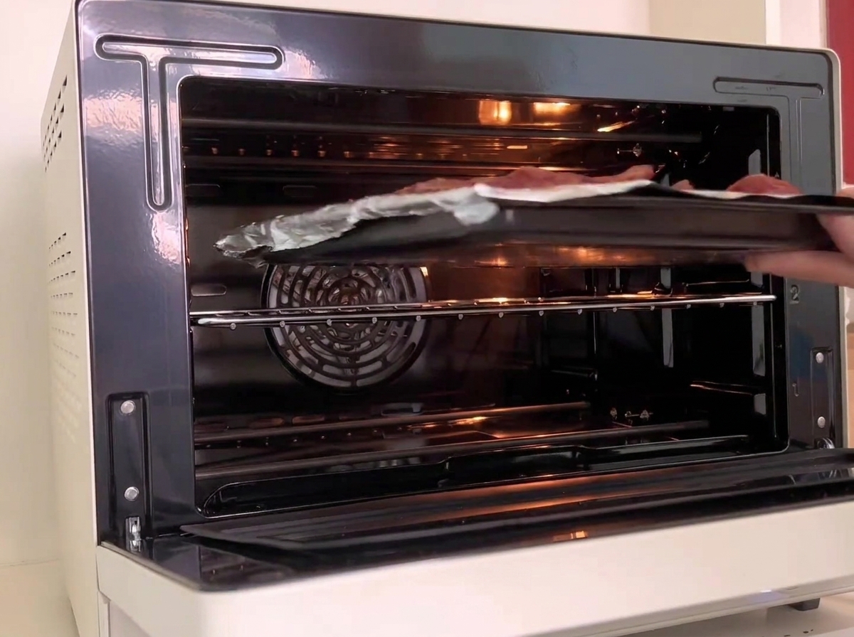 Un plat de cuisson contenant des côtes enveloppées de papier aluminium en train dêtre glissé sur la grille centrale dun four blanc éclairé.