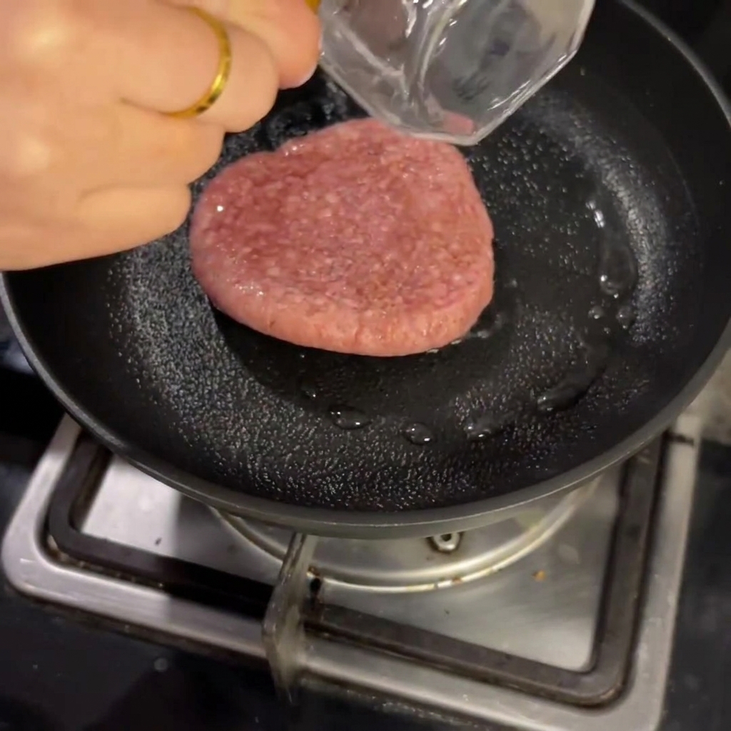 Un steak haché cru cuisant dans une poêle avec un peu deau, couvert dun couvercle.