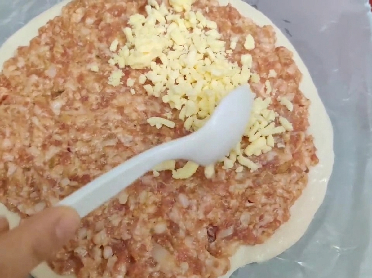 Une cuillère blanche étalant de la viande hachée crue et du fromage râpé sur une pâte ronde.