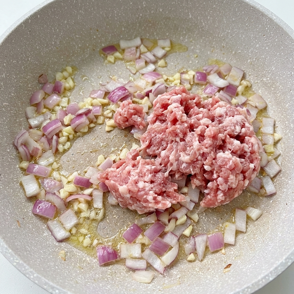 Porc haché cru dans une poêle à côté des oignons et de lail émincé en train de cuire.