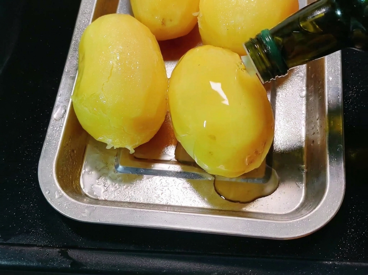 De lhuile de cuisson versée dune bouteille sur des pommes de terre épluchées dans un plateau de cuisson en métal.