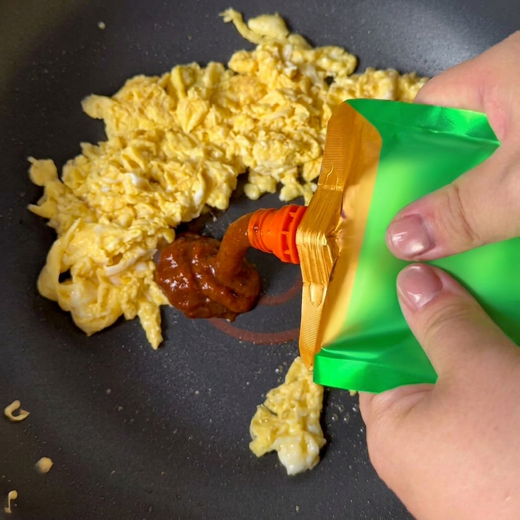 Pâte de soja jaune pressée dun sachet vert sur les œufs brouillés dans une poêle.