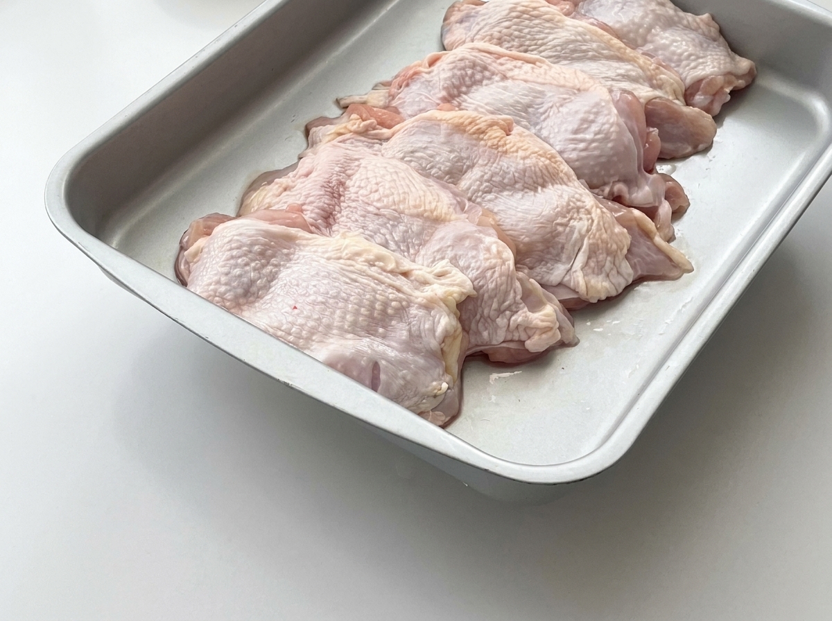 Cuisses de poulet crues disposées sur un plateau en métal avec des tranches de gingembre frais par-dessus.