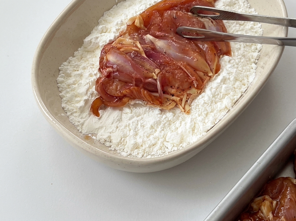 Un morceau de poulet mariné manipulé avec des pinces dans un bol rempli de fécule de maïs blanche.