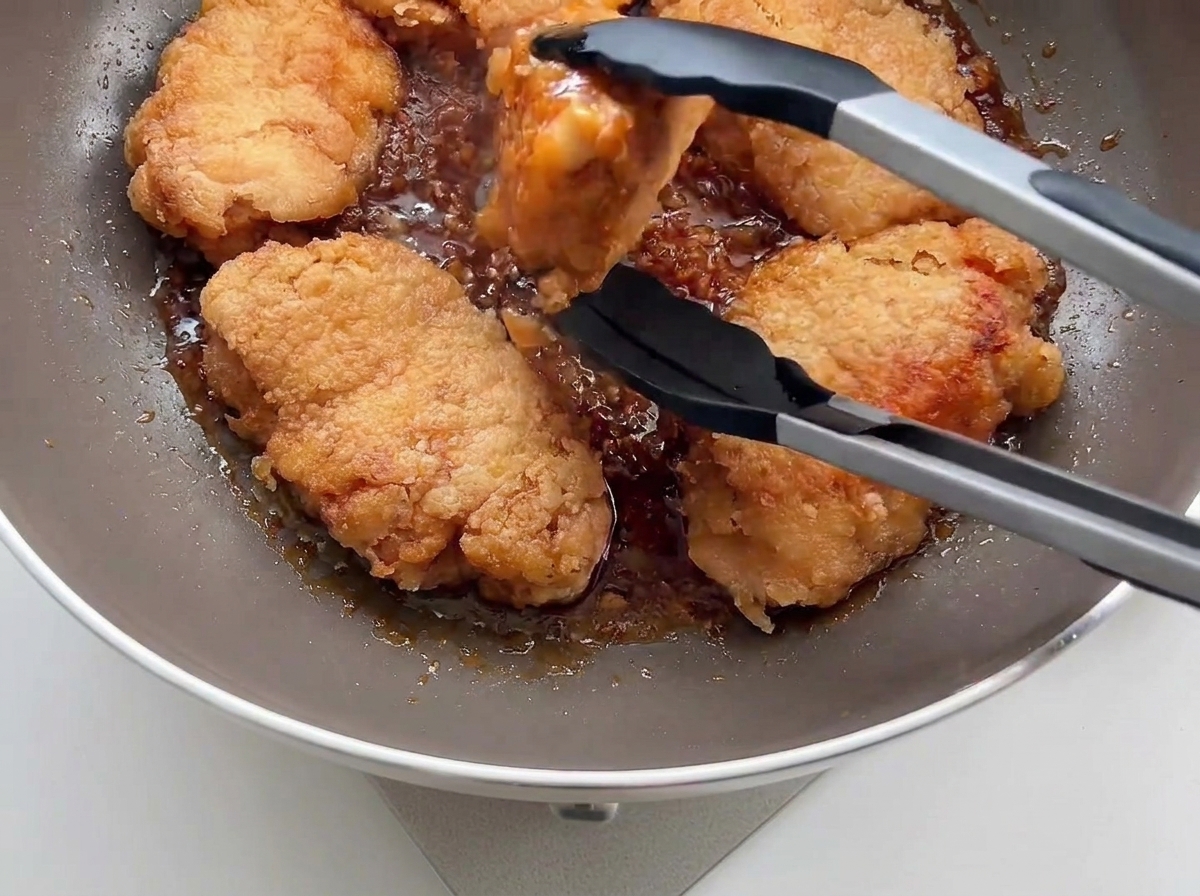 Poulet frit croustillant replacé dans une poêle avec la sauce à lail à laide de pinces de cuisine noires.