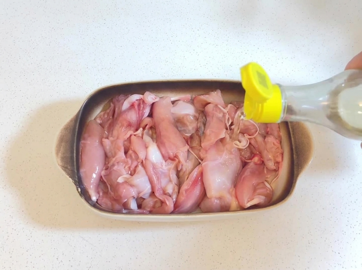 Du vin de cuisine transparent versé dune petite bouteille à bouchon jaune sur des morceaux de poulet crus dans un plat allant au four.