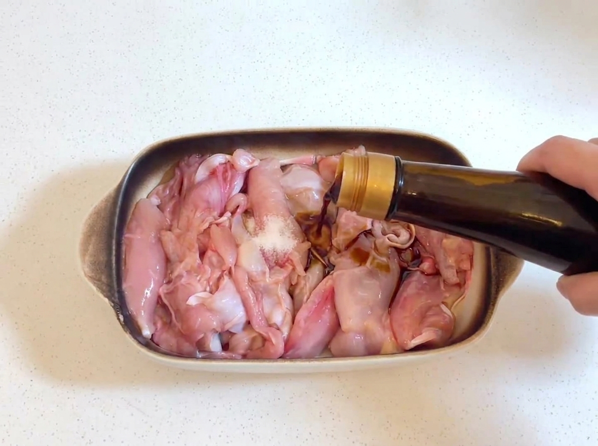 De la sauce soja foncée versée dune bouteille en verre sur des morceaux de poulet crus, avec une petite pincée de sel visible sur le poulet.