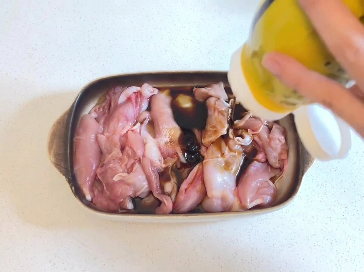 De la sauce huître épaisse et sombre pressée dune bouteille jaune sur des morceaux de poulet crus reposant dans une marinade à la sauce soja.