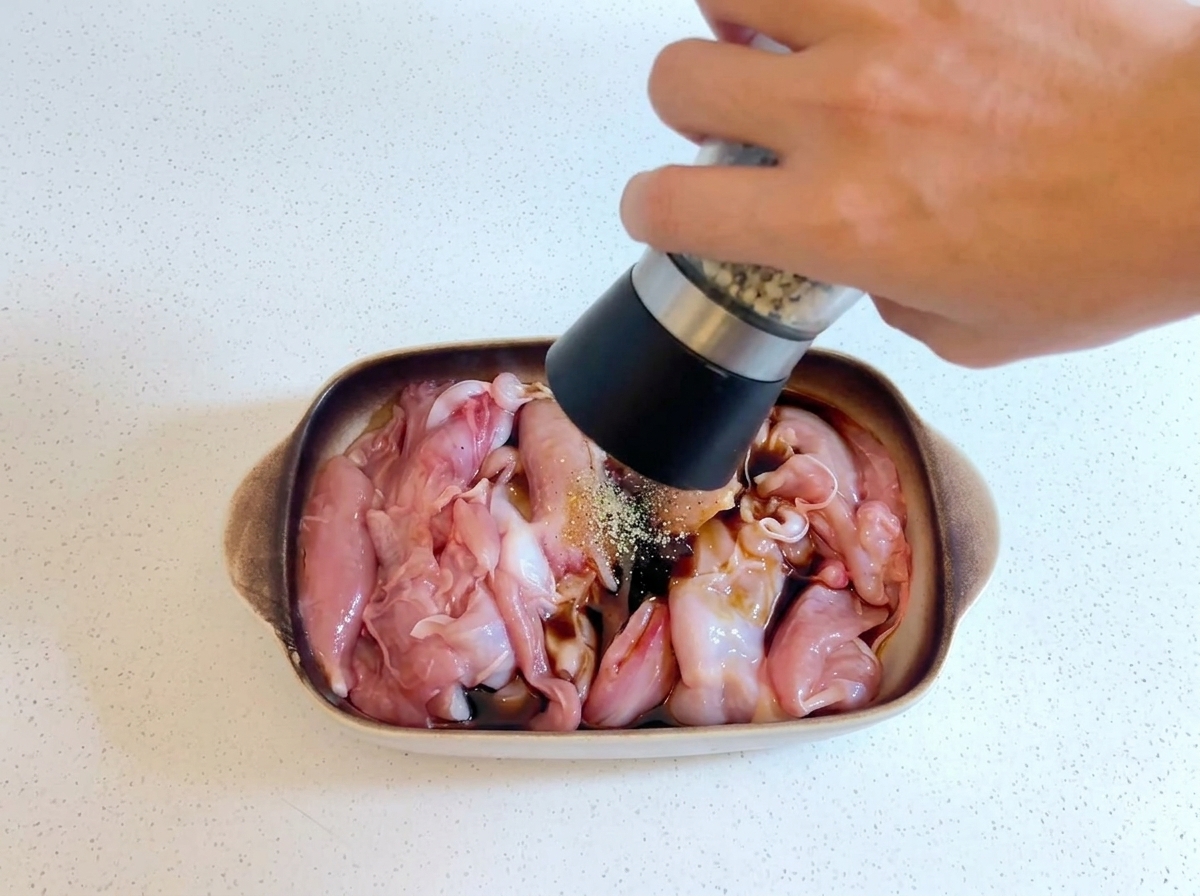 Une main tenant un moulin à poivre et moulant du poivre frais sur du poulet cru en train de mariner dans un plat.
