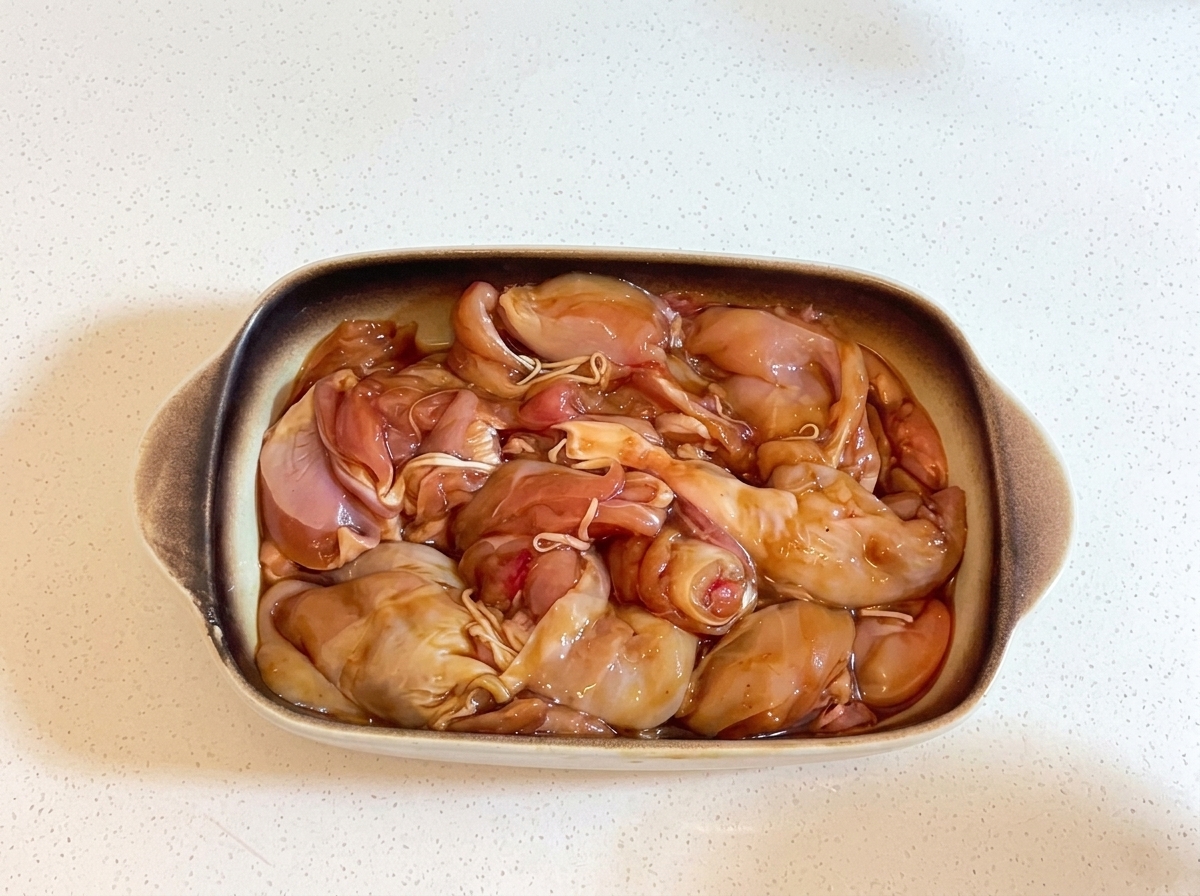 Un plat de cuisson rectangulaire en céramique rempli de morceaux de poulet marinés, hermétiquement recouvert de film plastique transparent.
