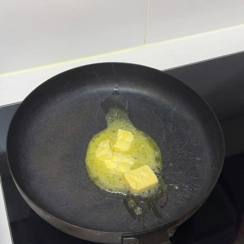 Des morceaux de beurre fondant en liquide dans une poêle sombre posée sur une plaque de cuisson.