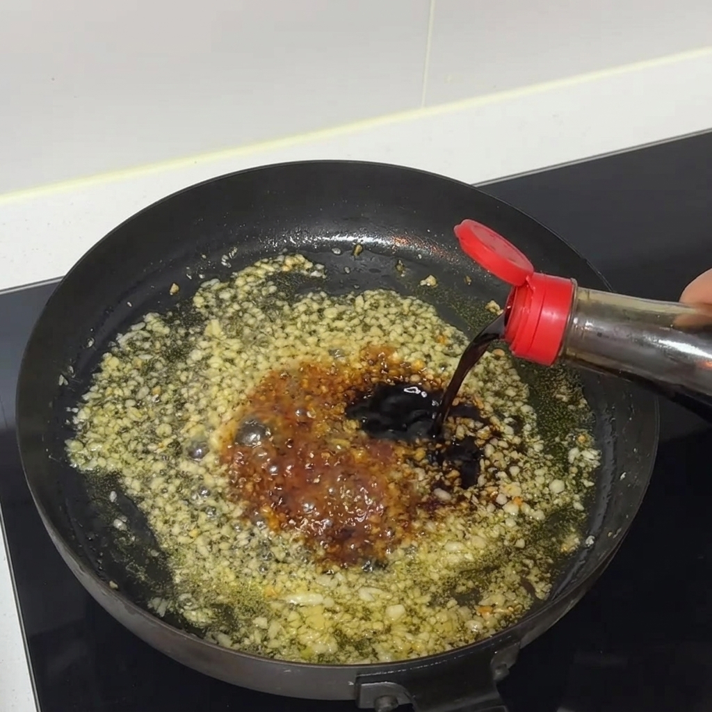 De la sauce soja noire versée dune bouteille dans une poêle grésillante contenant de lail haché et de la sauce.
