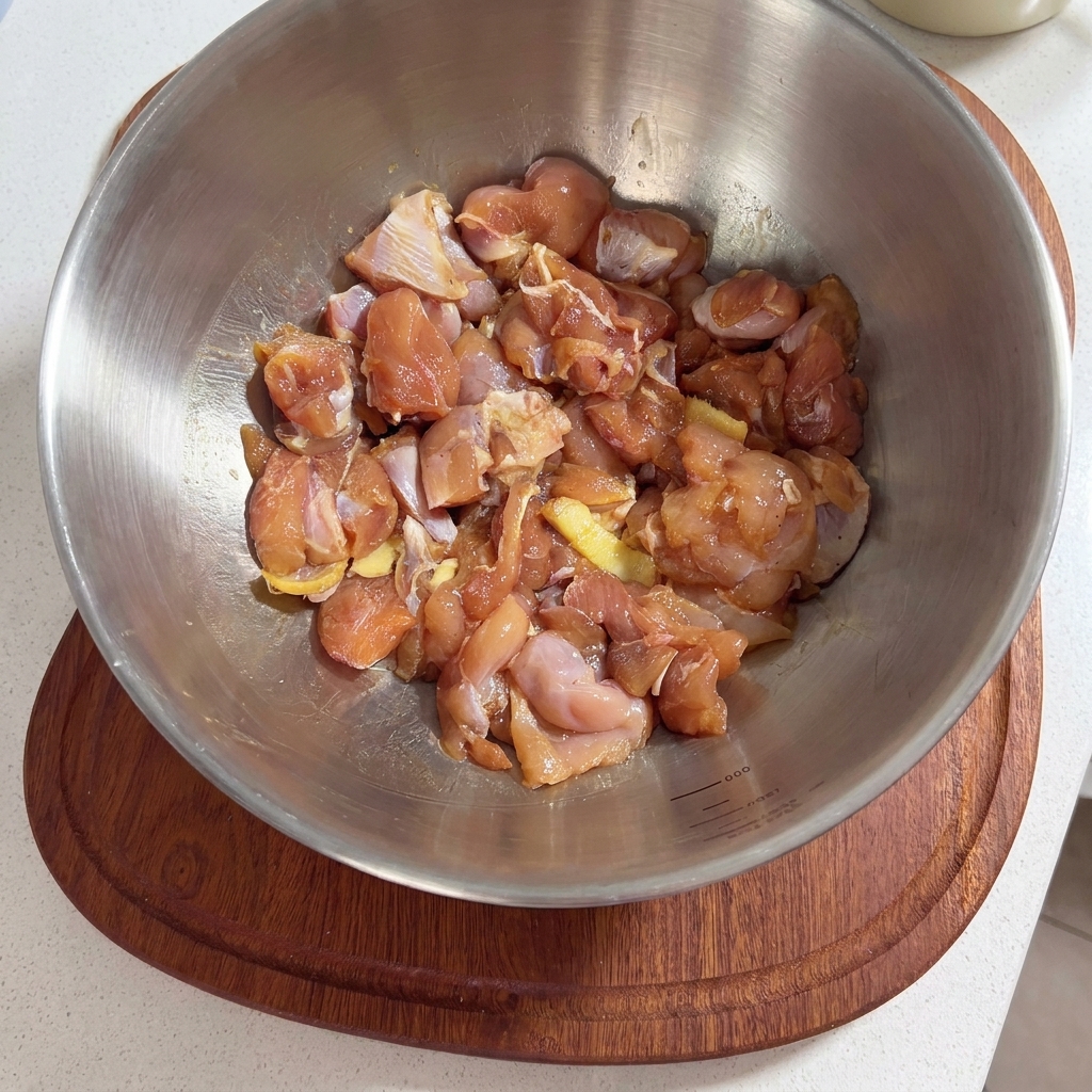 Un bol en métal rempli de morceaux de hauts de cuisse de poulet crus, assaisonnés de sauce de marinade et de tranches de gingembre.