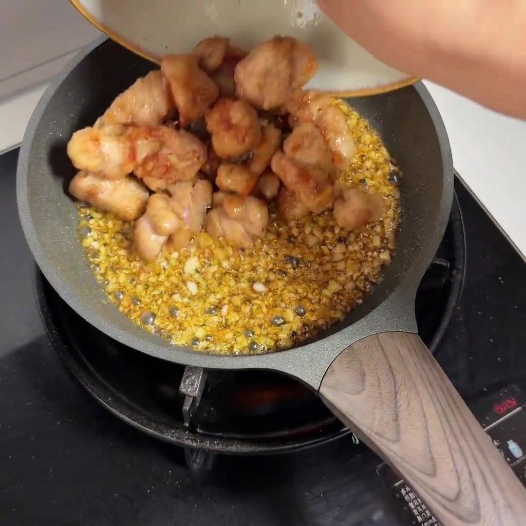 Morceaux de poulet pré-frits versés dune assiette dans une poêle remplie de sauce à lail.