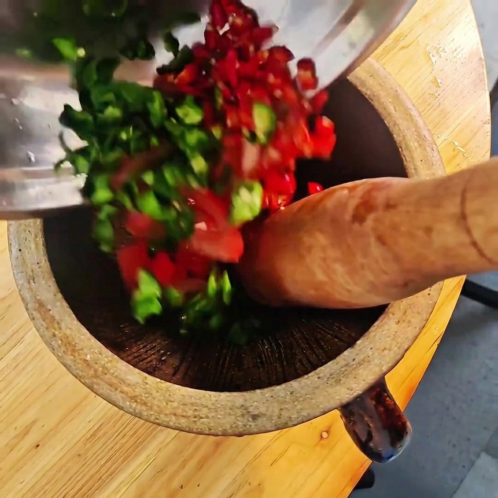Piments verts et rouges hachés et tomates en train dêtre écrasés dans un mortier en bois avec un pilon.