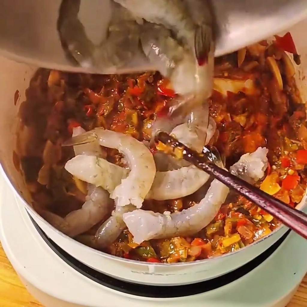 Crevettes entières grises crues jetées dans une sauce tomate rouge frémissante dans une casserole blanche.