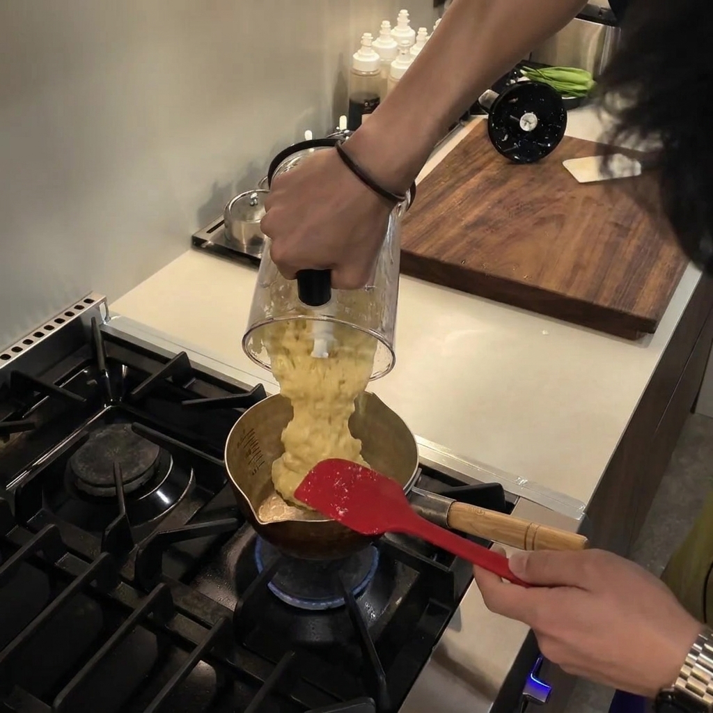Versement dune purée mélangée lisse et jaune pâle dun bol de mixeur vers une petite casserole métallique sur une cuisinière à gaz.