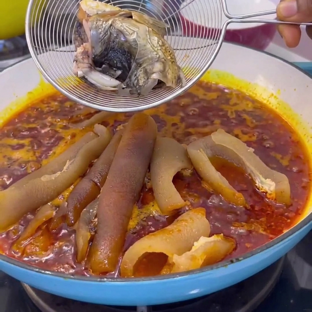 Une passoire métallique contenant des morceaux de poisson cuits descendue dans une casserole avec une sauce rouge frémissante et de la peau de porc.