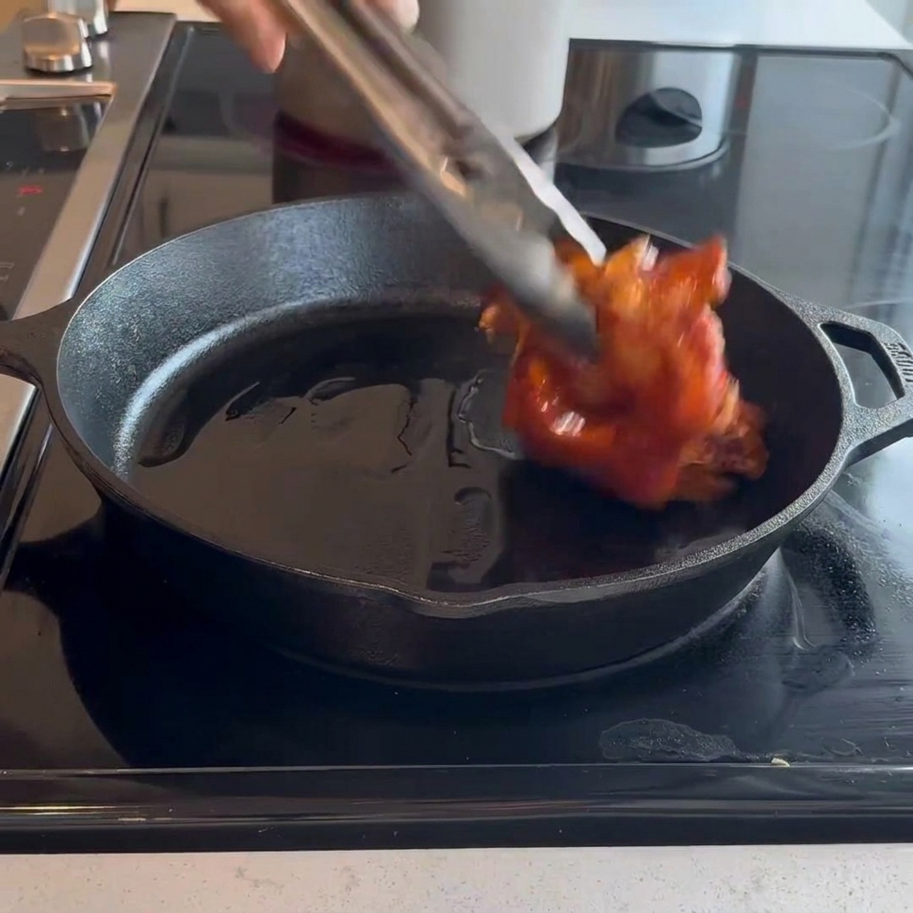 Des hauts de cuisse de poulet crus et marinés placés côté peau dans une poêle en fonte chaude à laide de pinces en métal.