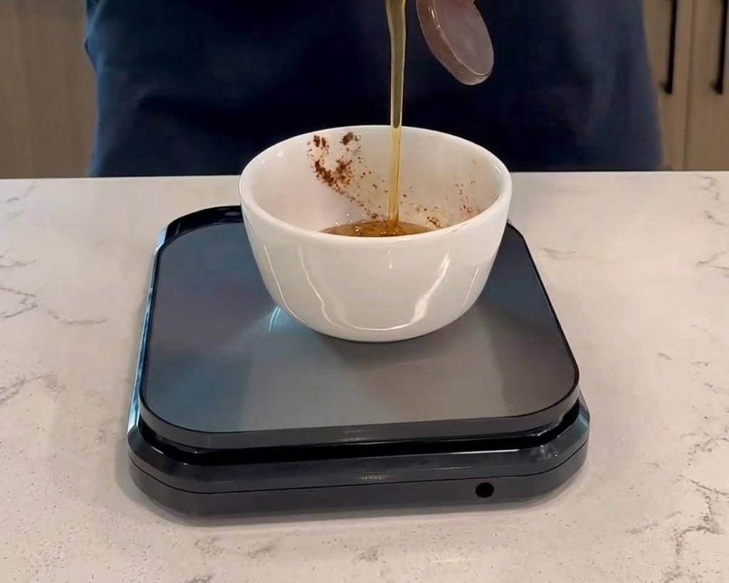 Verser du miel dune bouteille en plastique dans un petit bol blanc contenant de la sauce rouge foncée, posé sur une balance de cuisine numérique noire.