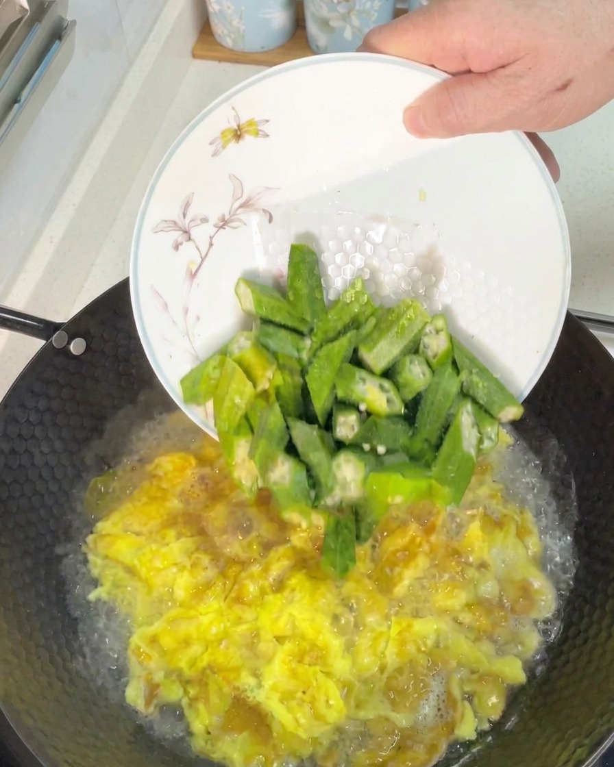 Ajout de morceaux de gombos verts fraîchement coupés dans un wok de soupe aux œufs bouillonnante.