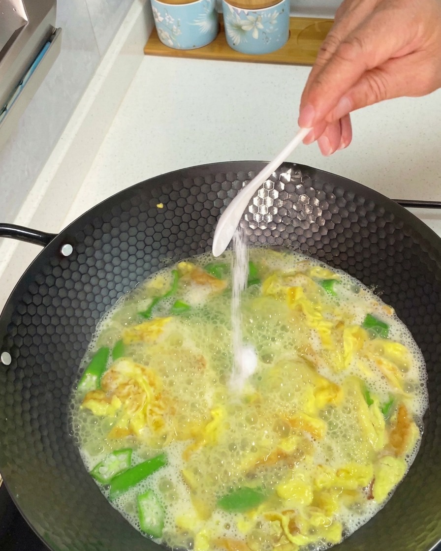 Ajout à la main dune cuillerée de sel blanc à un wok de soupe aux gombos et aux œufs frémissante.