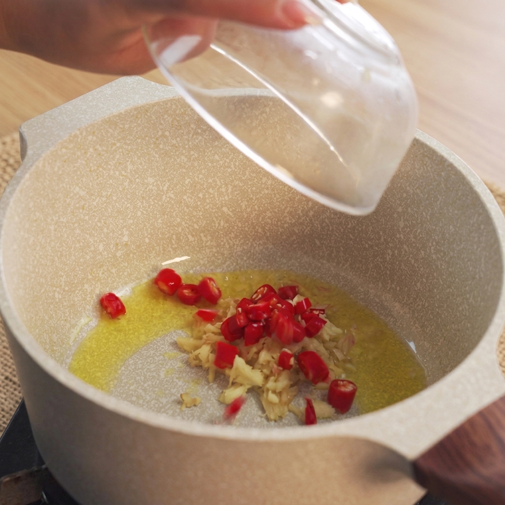 Une main jetant des piments rouges hachés dans une casserole contenant de lhuile chauffée et du gingembre émincé.