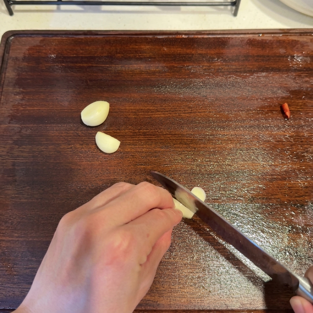 Des gousses dail coupées en rondelles avec un couteau sur une planche à découper en bois, à côté dun morceau de piment rouge.