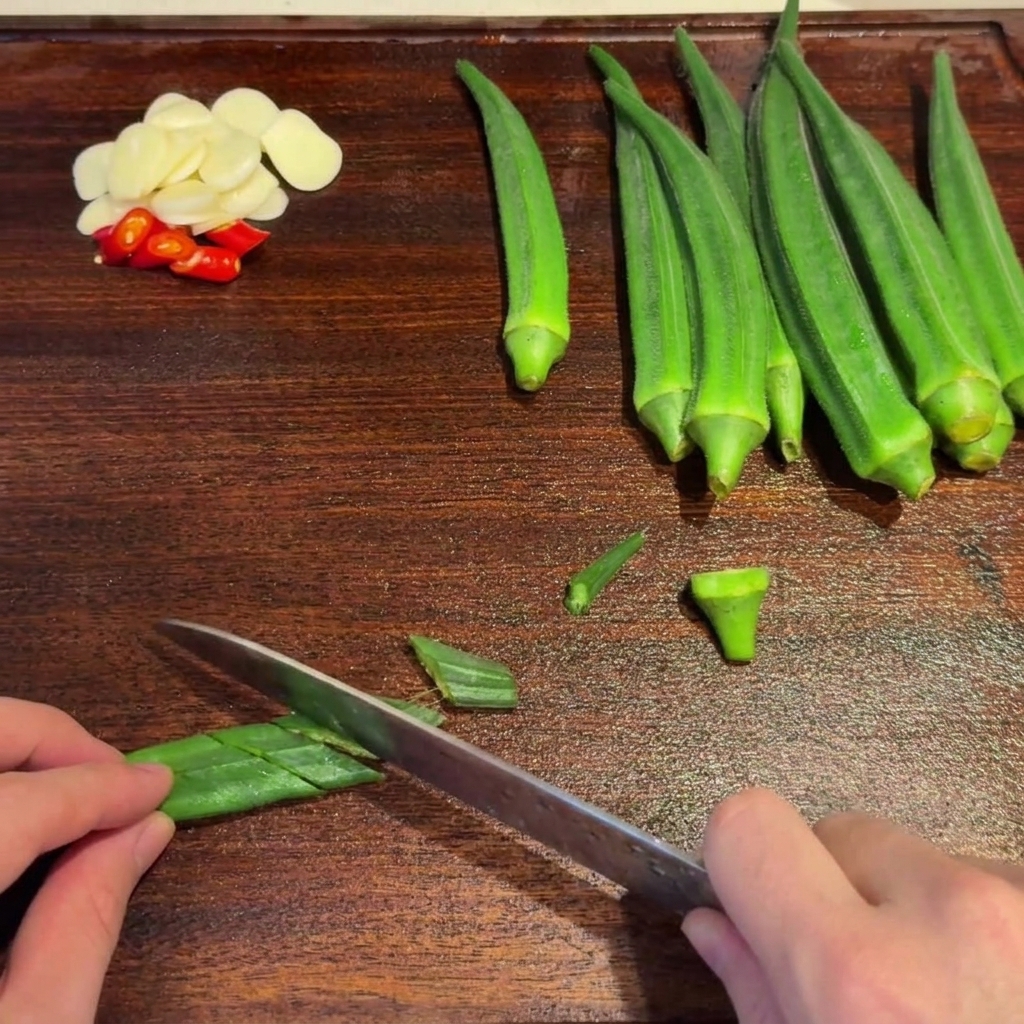Des gombos verts blanchis coupés en biseau avec un couteau sur une planche à découper en bois.