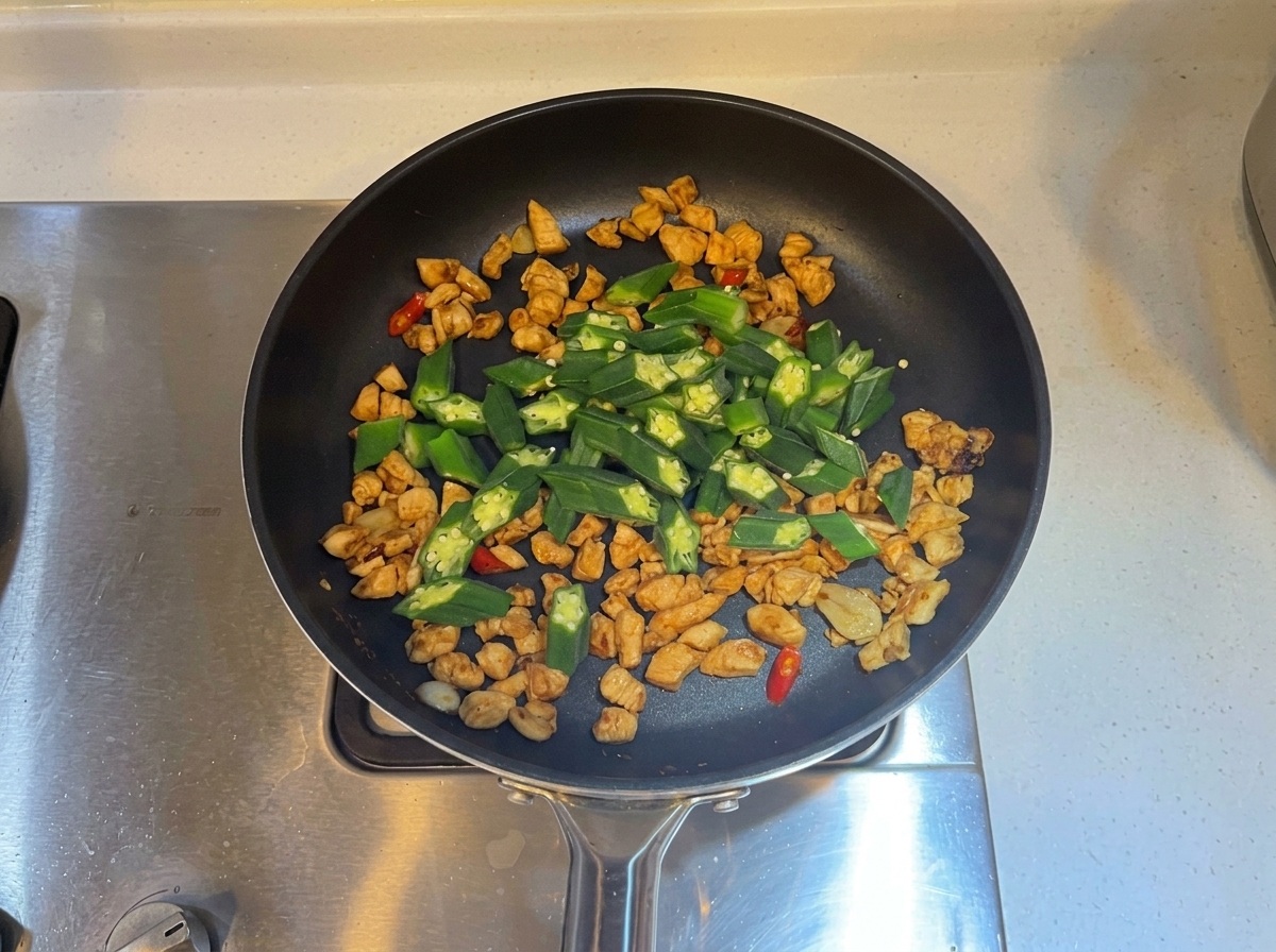 Des tranches de gombos verts mélangées dans la poêle avec le poulet cuit et les aromates.