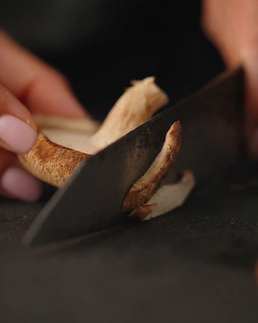 Gros plan sur des champignons frais en train dêtre émincés sur une planche à découper foncée avec un couteau.