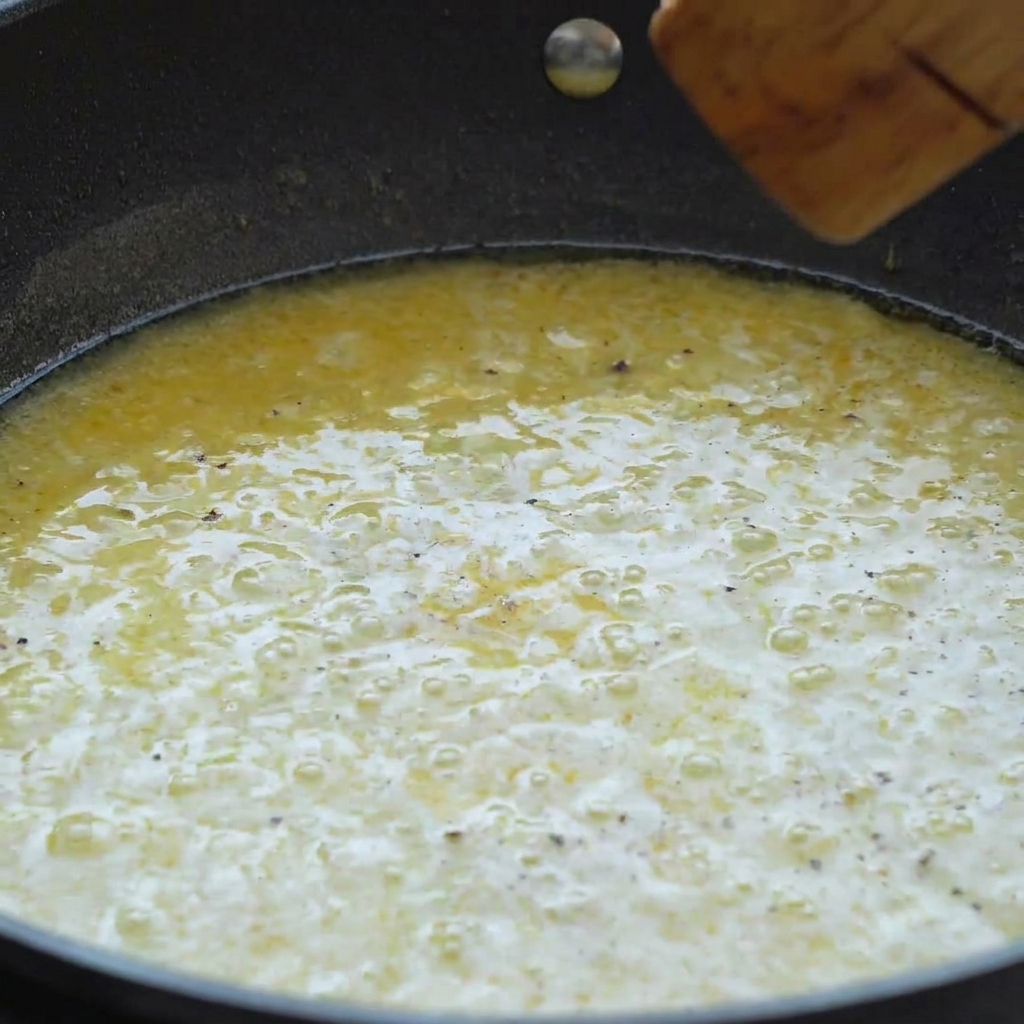 Une poêle remplie dune sauce crémeuse au beurre et au citron frémissante mélangée avec une spatule en bois.