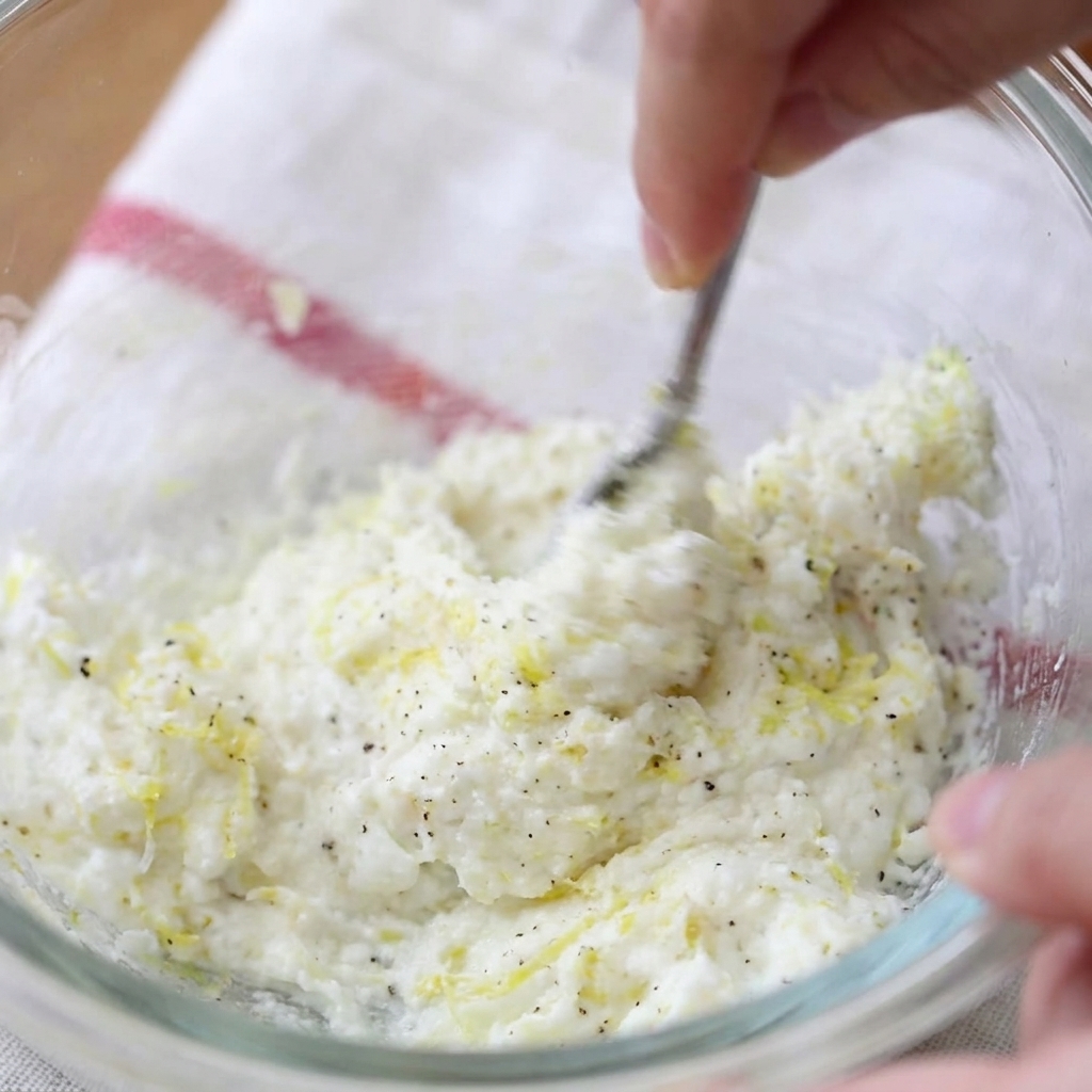 Une main utilisant une fourchette pour mélanger le fromage frais, le zeste de citron et le poivre noir dans un bol en verre.