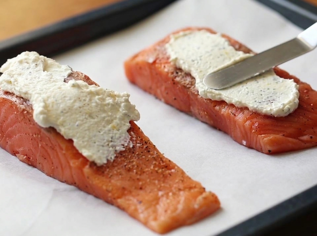 Une spatule en métal étalant une couche épaisse de mélange de fromage frais blanc sur deux filets de saumon crus posés sur une plaque de cuisson recouverte de papier sulfurisé.