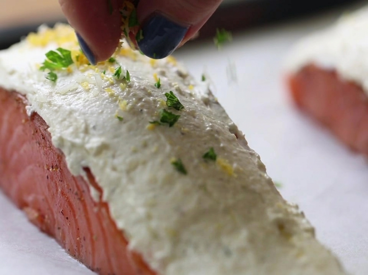 Des doigts saupoudrant un mélange de chapelure aux herbes vertes et jaunes sur des filets de saumon crus recouverts de fromage frais blanc.