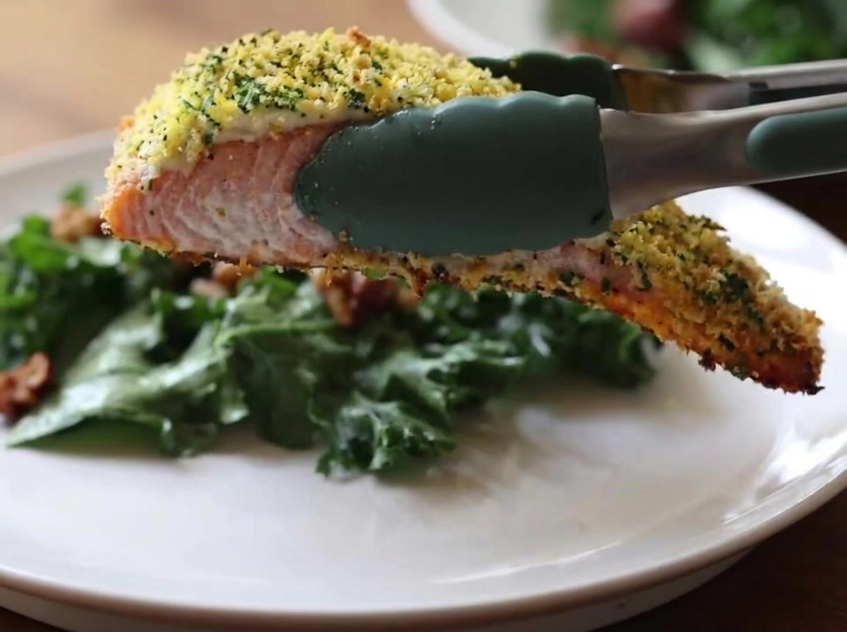 Des pinces de cuisine en silicone vert tenant un morceau de saumon cuit avec une croûte dorée et croustillante au-dessus dune assiette blanche contenant une salade de chou frisé vert.